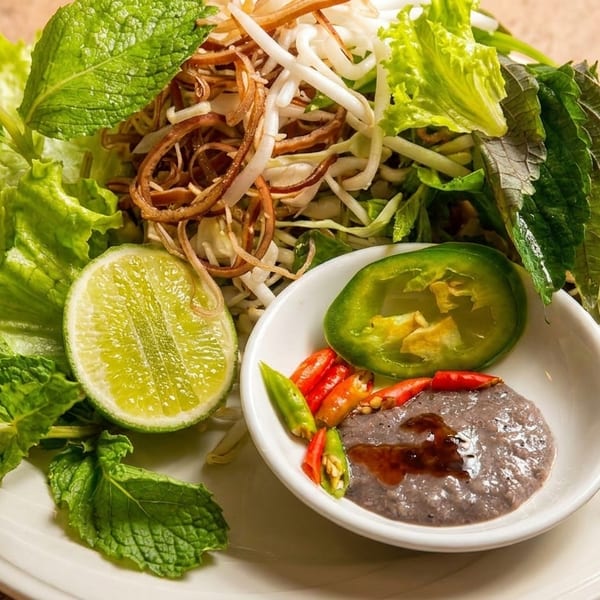 A vibrant plate of fresh herbs, lettuce, bean sprouts, lime, and a dipping sauce with chilies.