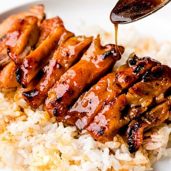Grilled teriyaki chicken slices with a glossy sauce being drizzled over white rice on a plate.